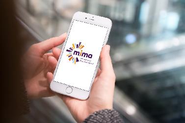 Eine Person hält ein Smartphone mit dem MIMA-Logo darauf. Die Umgebung ist modern und urban.
